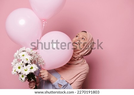 Happy Arab Muslim woman dressed in a pink hijab, rejoicing, experiencing happiness and amazement at a cute bouquet of white flowers and pink pastel helium balloons, isolated pink color background
