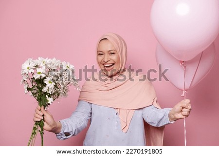 Happy Muslim woman in hijab, laughing looking at camera, posing with a bunch of white wild flowers and pink pastel helium balloons, on isolated background. Celebration. Congratulation. Festive events