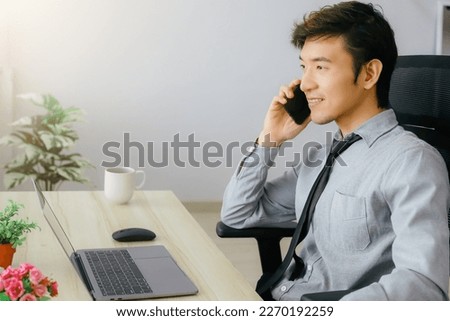 Asian businessman sitting at desk discussing business deal on mobile phone There are laptops and coffee mugs on the wooden table. Demonstrates success and confidence in leadership.