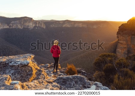 Golden sunlight streaming into valley with rising sandstone escarpment mountain and onto a cliff ledge where a female hiker standing taking in the magnificent views Royalty-Free Stock Photo #2270200015