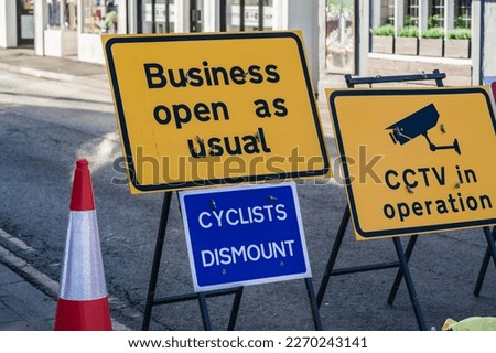 Business Open as usual, cyclists dismount, CCTV sign and traffic cone in front of road work background, concept illustration.