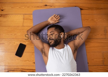 Young handsome black man resting on a yoga mat in between doing fitness exercises at home, listening to music in earphones