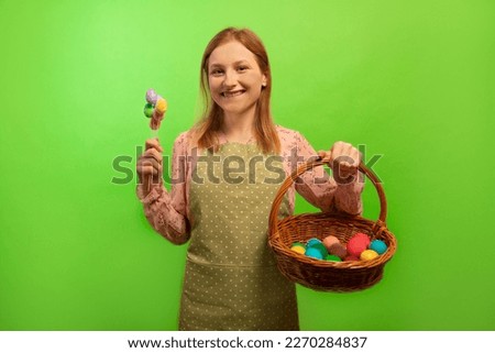 Happy smiling cheerful girl in green polka dot kitchen apron holding multicolored painted easter eggs on sticks and wicker basket with colored dyed easter eggs isolated on green background.