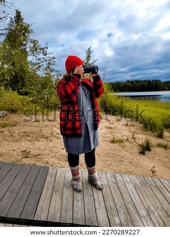 Young woman photographer at beach taking a photo. Beautiful lake landscape in the background.  Travel concept adventure active vacations outdoor. Woman wearing a red beanie hat and warm clothes.