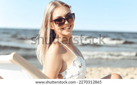 Happy blonde woman wearing sun glasses and relaxing on a wooden deck chair at the ocean beach.