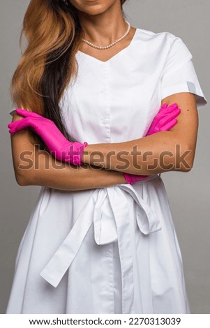 A woman in a white lab coat and pink latex gloves stands with her arms crossed against a light background. Medical staff