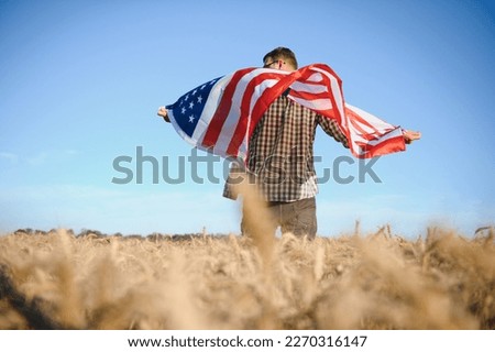Young patriotic farmer stands among new harvest. Boy walking with the american flag on the wheat field celebrating national independence day. 4th of July concept
