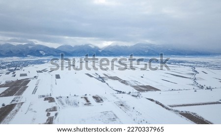 Aerial drone view over a small village located in an open field at the feet of Fagaras Mountains. Winter season - everything is snowed and frozen. Clouds are covering the sky.  Royalty-Free Stock Photo #2270337965