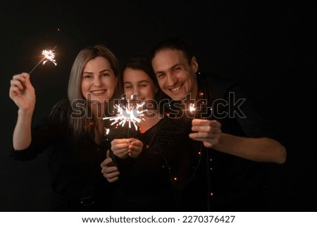 Happy family in black - woman, man and little girl, with a sparkler in hand the New Year's Christmas at home. The girl at the father on hands. New year 2023.