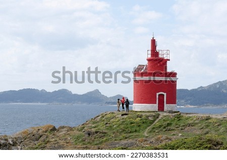 Tourists take pictures near the red lighthouse Punta Robaleira against the background of the sea and the island with high mountains. Travels in Spain. Concept of recreation with friends.