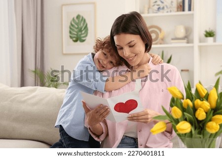 Little son congratulating his mom with Mother`s day at home. Woman holding handmade greeting card