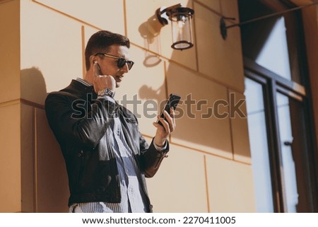 Young stylish fashionable man in black leather jacket shirt glasses stand near building wall outdoors, talk by mobile cell phone use air pods headphones Concept of people urban lifestyle and clothes.