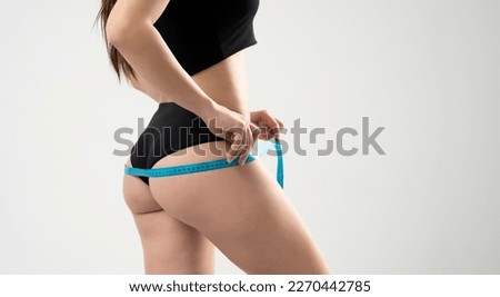 Young woman in black underwear is measuring her thigh with measuring tape on white background.