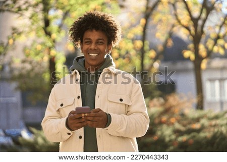 Cool smiling young African American guy holding mobile phone standing with smartphone at big city sunny street. Happy stylish hipster gen z teen using cellphone device outdoors, looking at camera.
