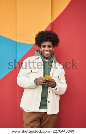 Happy smiling young African American hipster gen z guy standing at color bright red wall outdoors using cell phone, checking mobile, looking at camera holding cellphone. Vertical Royalty-Free Stock Photo #2270443349