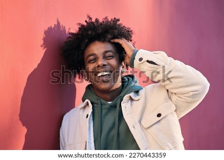 Young happy African American cool hipster guy face laughing on red city wall lit with sunlight. Smiling stylish cool rebel gen z teenager model standing outdoors. Headshot close up portrait shot.