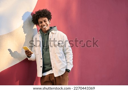 Happy cool gen z African American ethnic stylish hipster guy model standing at red city urban wall outdoors lit with sunlight using cell phone mobile device, looking at camera holding cellphone.