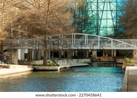Metal bridge over canal or lake in urban or commercial district in mall or outdoor shopping center in san antonio texas downtown. Reflective glass building in afternoon sun with sidewalks.
