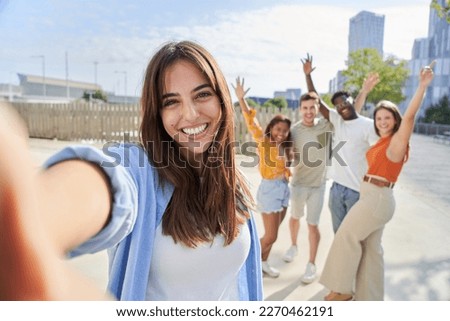 POV selfie outside of a smiling and cheerful pretty young girl with her friends in the background. Happy people looking camera and celebrating with hands up. Multiracial portrait college classmates.