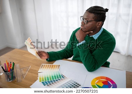 Pensive African American Male Designer Choosing Color Scheme Holding Palettes Sitting In Modern Office. Professional Design Career And Colors Trends Concept. Side View
