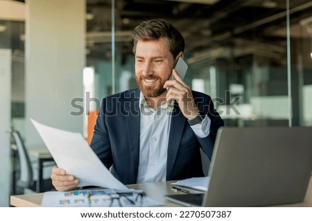 Corporate communication. Happy male manager working remotely, reading financial document and talking on mobile phone, sitting at desk in office interior