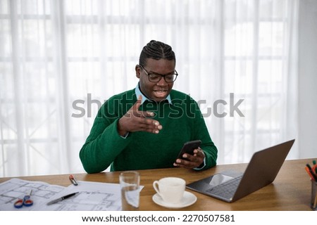 African American Businessman Using Voice Search On Smartphone Working Online Via Laptop Computer Sitting In Modern Office. Business Communication And Technology Concept
