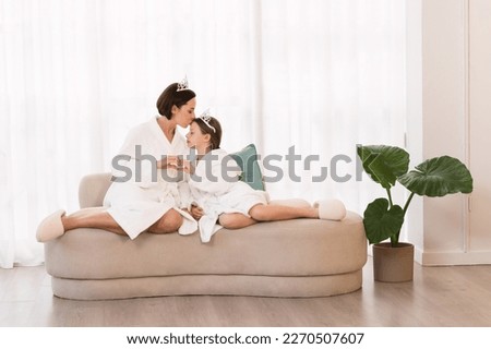 Loving Mother And Little Daughter Wearing Bathrobes And Crowns Bonding At Home, Beautiful Mom And Female Child Making Heart Sign With Hands, Relaxing On Couch After Spa Day, Mommy Kissing Girl