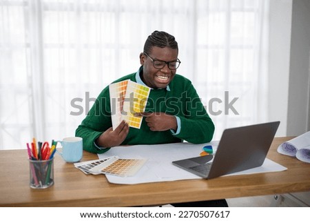 Smiling Black Designer Man Making Video Call Showing Palette To Laptop Computer Choosing Color Scheme With Client Sitting At Workplace Indoors. Successful Design Career Concept