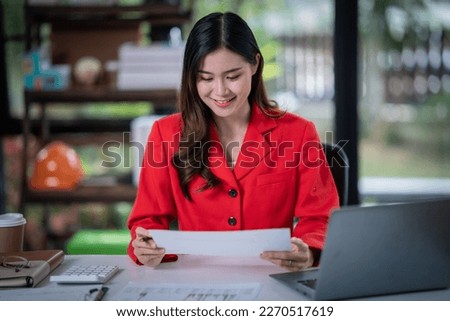 Attractive young smiling Asain woman in smart casual wear and holding smart phone looking at camera while leaning on desk in creative office Confident business exper.