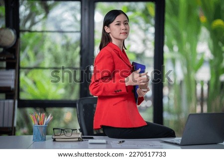 Attractive young smiling Asain woman in smart casual wear and holding smart phone looking at camera while leaning on desk in creative office Confident business exper.