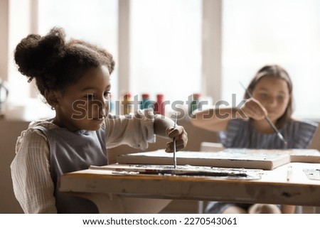 Focused pretty little pupil girl in apron taking lesson in creative studio school, painting on canvas at table, using paintbrush and palette, training artistic talent, drawing skills