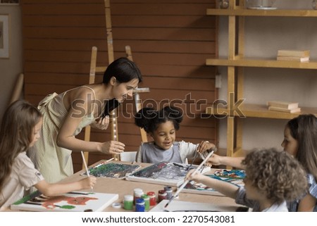 Friendly young artistic class teacher helping pupil girl with drawing on canvas, discussing picture with African artist kid, smiling, laughing. Group of children learning painting in artistic school