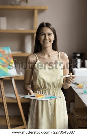 Happy inspired pretty artistic school teacher using palette and paintbrush, posing in creative studio with easel and picture in background, enjoying craft creativity, craft hobby. Vertical portrait