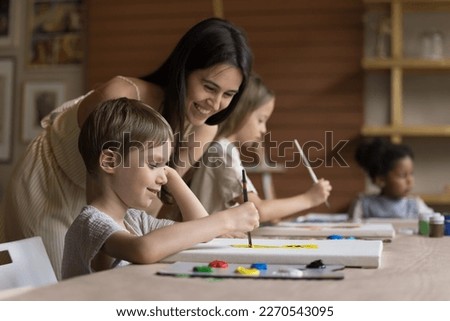 Cheerful pretty teacher girl helping pupil boy with drawing picture on canvas, standing at kid, smiling, laughing, looking at canvas. Child attending artistic school class, learning painting