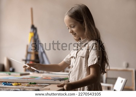 Happy little kid enjoying creative hobby, studying painting in artist school, drawing picture at big table in craft studio, mixing paints on palette, smiling, training art skills