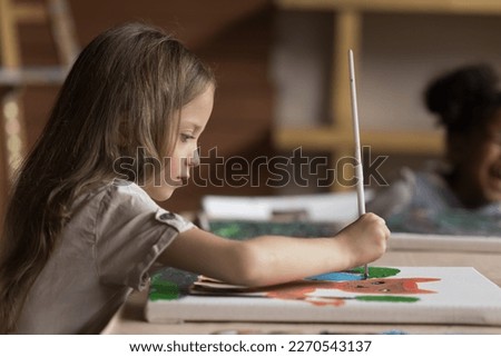 Focused cute pretty art school pupil girl drawing cartoon animal on canvas, studying creativity on painting class, holding paintbrush, sitting at big table in classroom. Side view
