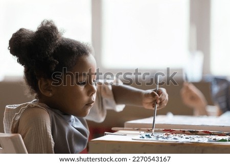 Serious artistic school African American pupil girl painting on canvas on group class, mixing paints on palette with paintbrush, studying creativity, working at table in studio
