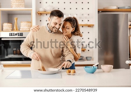 A happy man is stirring eggs in pan in kitchen at home while his wife is hugging him.