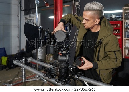 handsome caucasian man with hair videographer changing the lens on a professional movie camera that stands on a camera trolley on rails on a film set