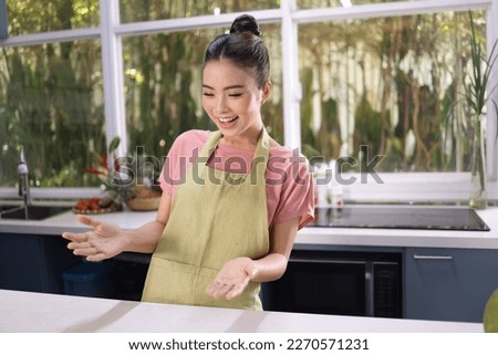 Portrait of a young beautiful pretty cheerful Asian young lady, wear apron, in the kitchen with hand gesture to show the surprise things on copy space Royalty-Free Stock Photo #2270571231