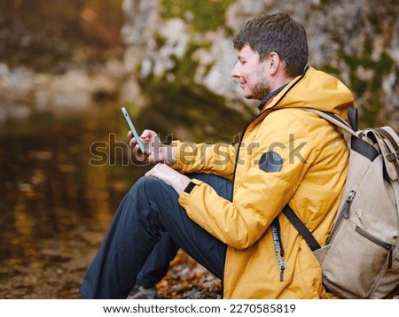 Adventure and active lifestyle in nature. Tourist hiking in forest. Caucasian man in yellow jacket walks in woods. using navigation application on smartphone, social networks, taking pictures
