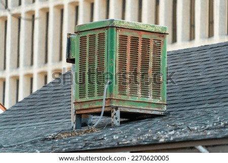 Swamp cooler air condition system on rooftop in modern city with green color and aged and weathered metal body. Gray roof tiles with white cement building background in afternoon shade. Royalty-Free Stock Photo #2270620005