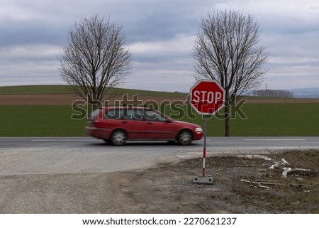 Crossroads with a stop sign as seen from our side. A red car is driving through from right to left. The surroundings show a cloudy landscape with two trees behind the car and fields in the background.