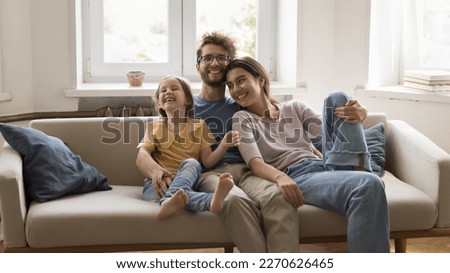 Happy handsome loving caring husband hugging cute laughing daughter girl and cheerful pretty wife, resting on home couch, relaxing in living room, enjoying family leisure time