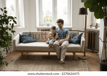 Positive young father and little daughter kid using mobile phone on home sofa for video call. Dad watching kid playing online game, using learning app for school study home task