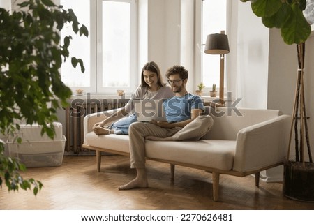 Cheerful millennial young husband and wife using laptop for online communication together, resting on couch, holding computer, talking on video call, watching TV, movie on Internet