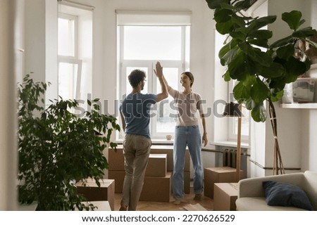 Positive young married couple clapping hands, giving high five over heap of paper relocation boxes in new flat, moving into eco decorated apartment with plants, celebrating property buying Royalty-Free Stock Photo #2270626529