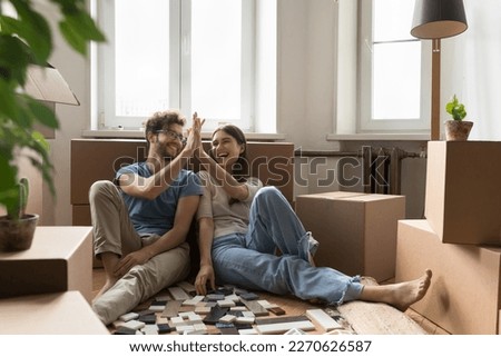 Cheerful excited young couple planning renovation after moving into new apartment, choosing interior material samples of tile, laughing, giving high five, clapping hands at cardboard boxes Royalty-Free Stock Photo #2270626587