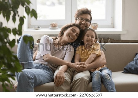 Cheerful attractive couple of young parents hugging cute girl on sofa, looking at camera, smiling, laughing. Family portrait of happy mom, dad and daughter kid enjoying closeness, leisure at home
