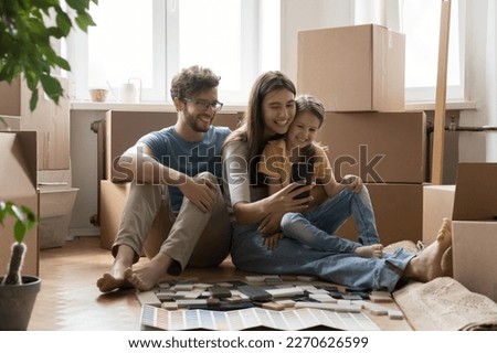 Positive parents and happy kids taking moving selfie on smartphone, sitting at stacked cardboard boxes, renovation brochures with wall color samples, holding mobile phone, smiling Royalty-Free Stock Photo #2270626599